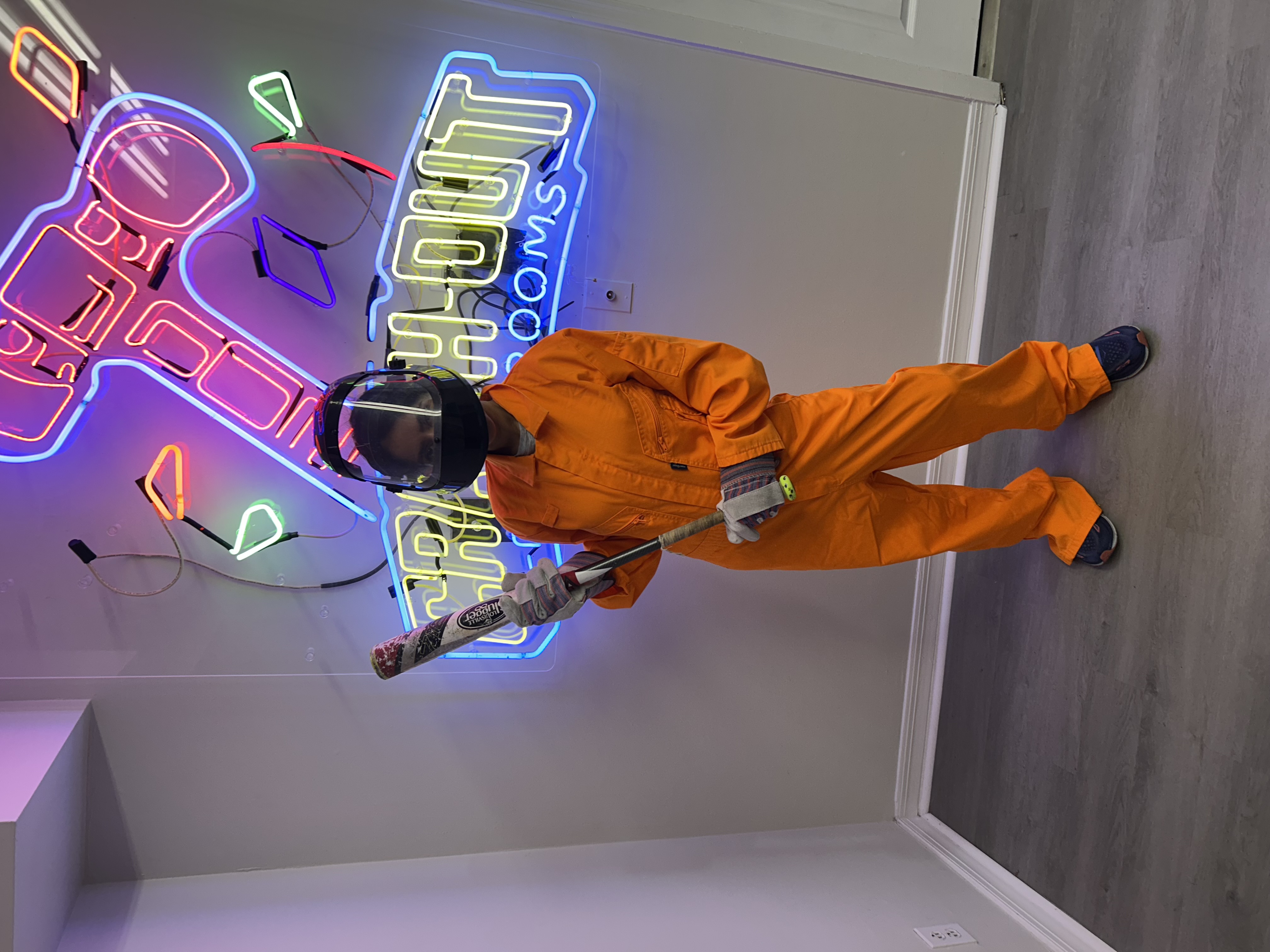 Crash Out customer in orange jumpsuit posing with baseball bat in front of vibrant Crash Out neon sign