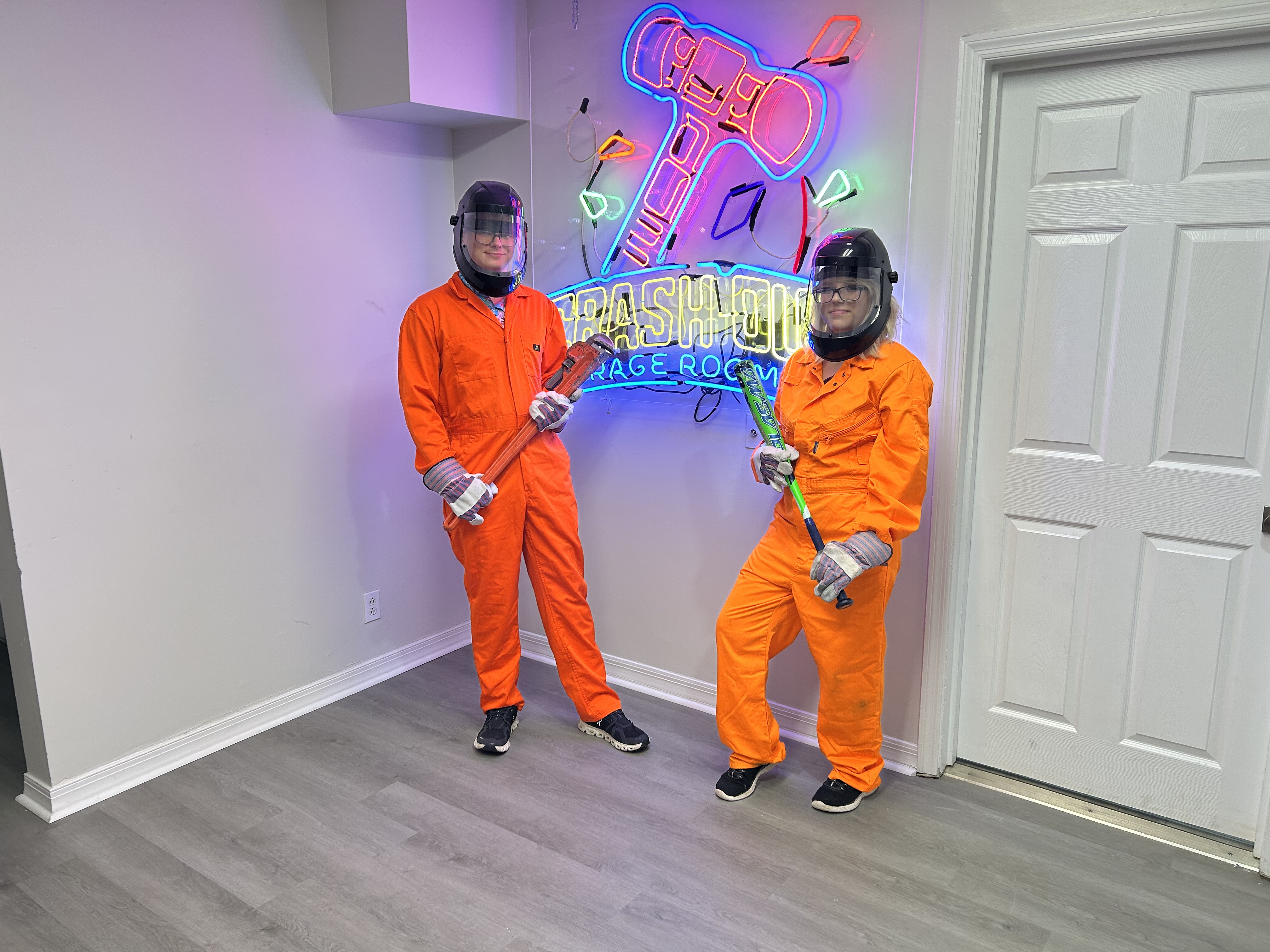 Large group of Crash Out Rage Rooms customers in orange jumpsuits holding bats and tools in front of branded neon sign